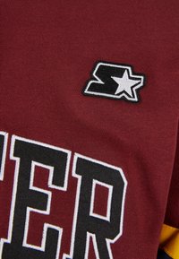 Maillot bordeaux avec de grandes lettres texturées en noir et blanc, agrémenté d'un petit logo brodé représentant un "S" et une étoile.
