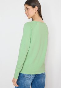 Femme aux cheveux bruns portant un pull vert clair à manches longues et un jean bleu, légèrement tournée sur le côté contre un fond blanc.