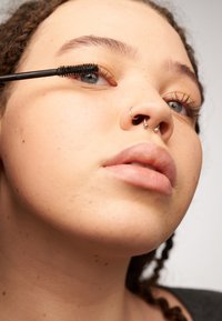 Brosse de mascara noire appliquant le produit sur les cils. Gros plan sur un visage à la peau claire, aux yeux bleus et avec plusieurs piercings faciaux.