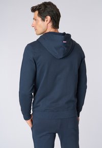 Felpa con cappuccio blu navy, con retro intero e polsini a coste. Presenta un piccolo logo sulla parte del collo e un tessuto liscio e texturizzato.