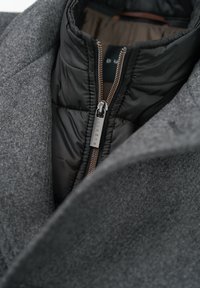 Grau gestalteter Wollmischungs-Mantel mit einem schwarzen, gesteppten Innenfutter; verfügt über einen hohen Kragen und bronzefarbenen Reißverschluss; glatte Textur mit strukturiertem Design.