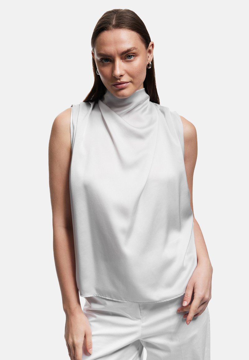 Mouwloze witte top van gedrapeerd, soepel materiaal, met hoge halslijn en een relaxte pasvorm. Gecombineerd met witte broek met minimale naden en strakke randen.