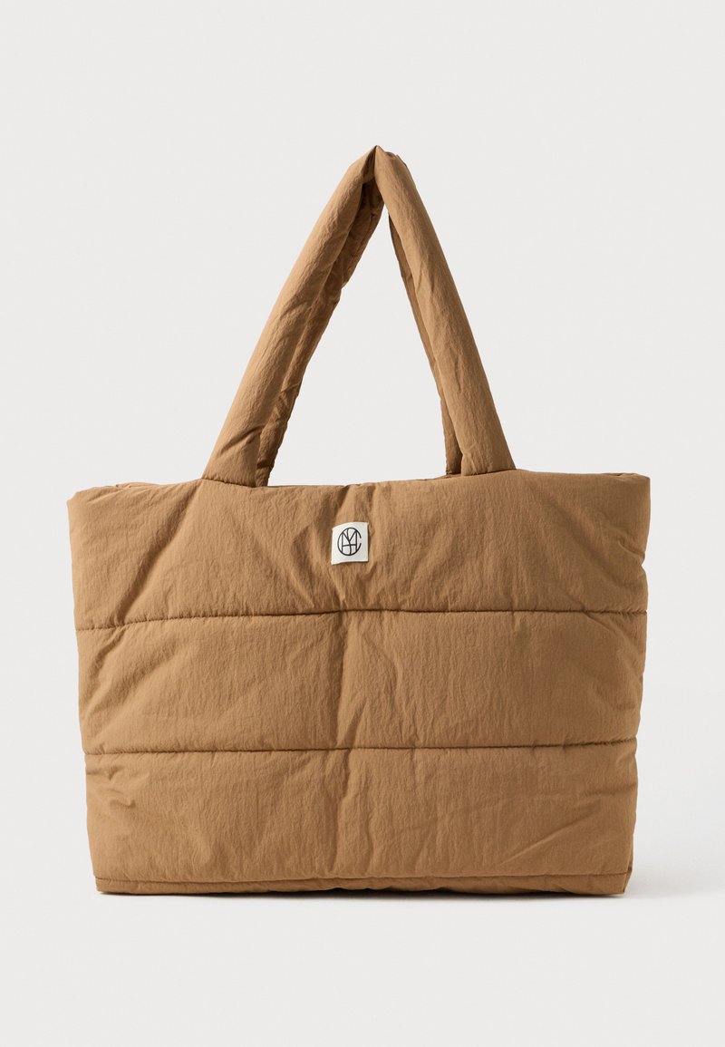 Brun polstret totebag med brede håndtak og en liten hvit etikett med svart sirkulært logo i midten foran.