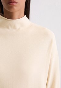Crème hoge nek sweater gemaakt van zachte, lichte stof met een gladde textuur en een aansluitend ontwerp dat tot de kraag reikt.
