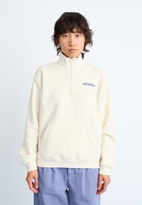 Sweatshirt crème, à col montant, avec une poche à l'avant et un petit logo "new balance". Tissu doux ; coupe décontractée et manches longues.