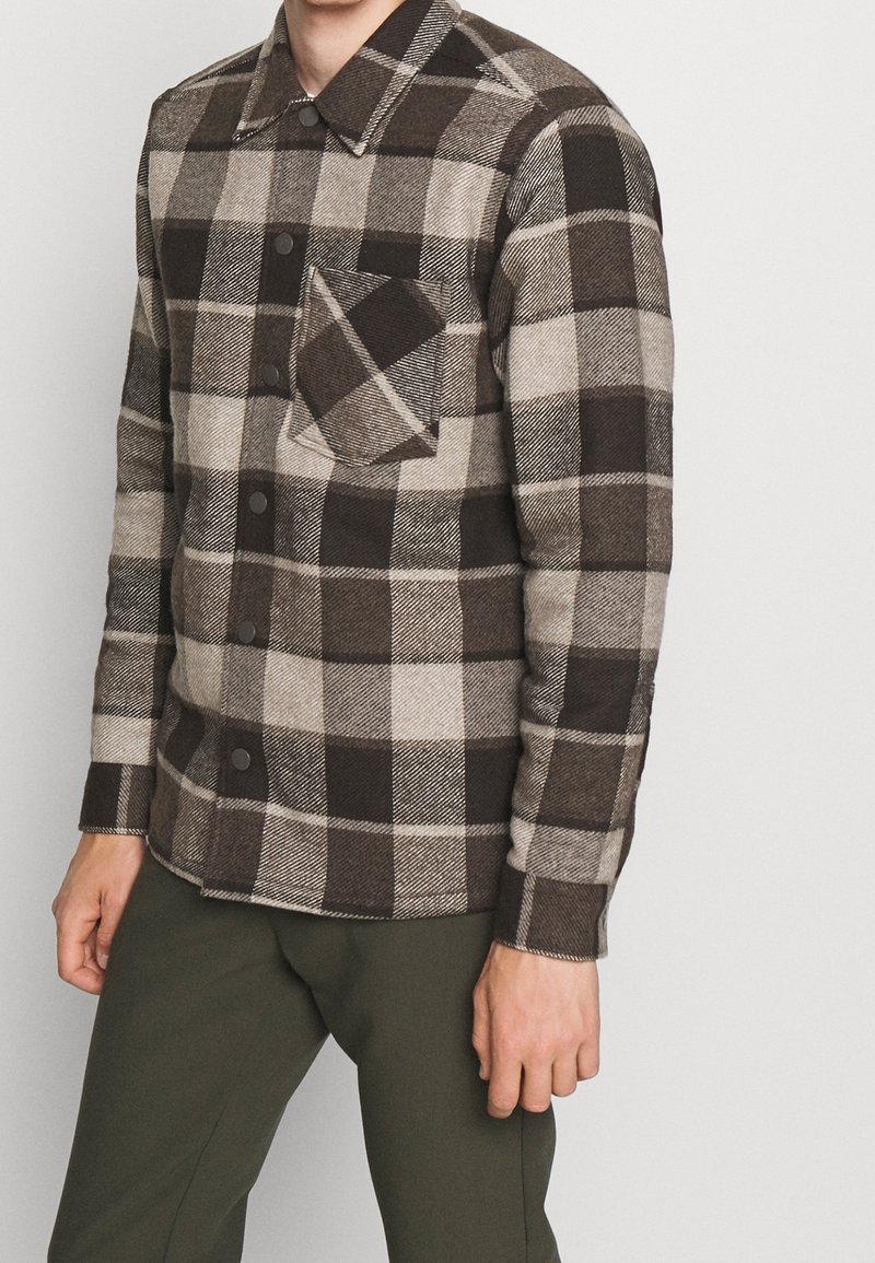 Homme portant une chemise à carreaux marron, beige et noire à boutons avec une seule poche poitrine et un pantalon vert olive, sur un fond clair uni.