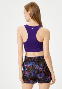 Lilla atletisk crop top med racerback-design og logo; parret med sorte shorts med et levende blomstermønster i blå og rød.