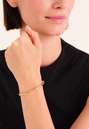 Gouden metalen armband met een bamboe-geïnspireerd ontwerp, met gladde, cilindrische schakels. Draagt op een pols tegen een donkere kledingachtergrond.