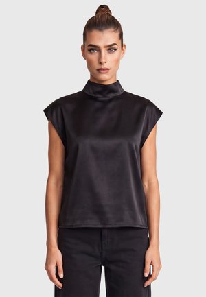 Frau mit hochgestecktem Haar trägt schwarzen Satin ärmellosen Top mit hohem Kragen und schwarze Hose, stehend vor hellgrauem Hintergrund.