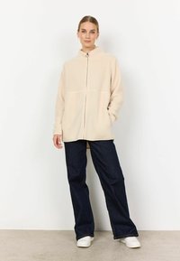 Beige fleece takki, jossa on korkea kaulus ja etuvetoketju, rento malli. Yhdistetty tummansiniset leveälahkeiset farkut ja valkoiset lenkkarit.