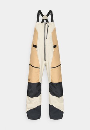 Mono de nieve ligero en crema, beige y marino; cuenta con tirantes ajustables, bolsillos con cremallera y una tela texturizada para mayor durabilidad.