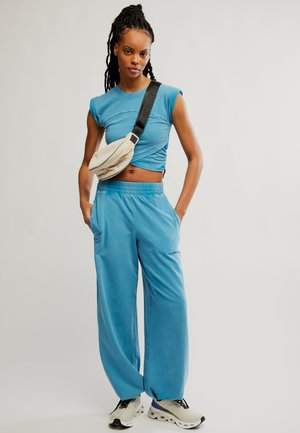 Junge Frau mit Dreadlocks, die ein blaues ärmelloses Oberteil, lockere blaue Hose, weiße Sneakers und eine beige Umhängetasche trägt.