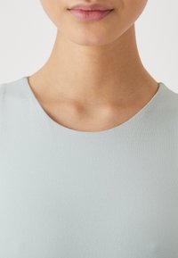 Close-up of a woman's lower face, neck, and upper chest wearing a light blue crew neck top against a plain background.