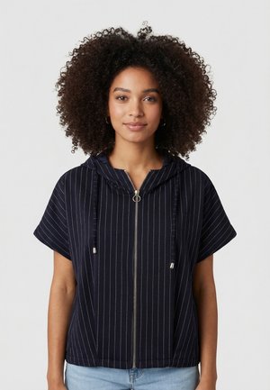 Mujer con cabello rizado que lleva una sudadera con capucha con cremallera de manga corta, azul marino con rayas finas, y jeans azul claro, posando contra un fondo liso.