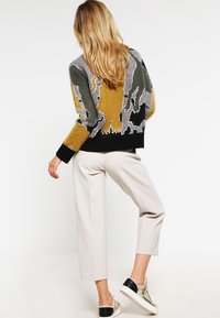 Bunter Pullover mit abstrakten Mustern in Grau, Grün und Senfgelb, schwarze Bündchen, kombiniert mit beigen, gekürzten Hosen und Sneakers.