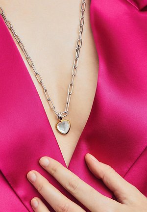 Zilveren ketting met een grote hartvormige hanger met een parelmoer inleg, gedragen tegen een roze satijnen top.