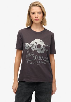 Lyshåret kvinde iført en sort t-shirt med en stor dødningehoved, rose og tekst, der blandt andet inkluderer "The 1010 Club" og japanske tegn.