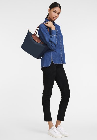 Navy tote bag with brown leather accents, paired with a denim shirt and black pants, worn by a person standing with a casual pose.