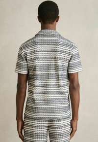 Set de cămașă cu mânecă scurtă și pantaloni scurți, fabricat dintr-un material alb texturat, cu dungi orizontale bleumarin și modele geometrice repetitive.
