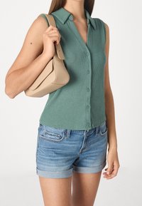 Vihreä hihaton paita, jossa on napit, yhdistetty vaaleansinisiin pöyheisiin farkkushortseihin. Oikeassa kädessä pidetään ruskeaa, jämäkkää käsilaukkua.