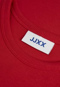 Červená ryhľovaná tkanina s okrúhlym výstrihom. Na oblasti goliera je viditeľná biela etiketa s modrým textom "JJXX".