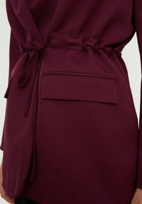 Veste bordeaux avec une taille cintrée, dotée d'un cordon de serrage et d'une poche à rabat. Texture lisse et lignes épurées qui valorisent le design.