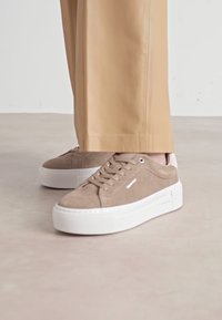 Beige Wildlederschuhe mit einer Plattform aus weißem Gummi, Schnürdesign und dezentem Logo-Detail an der Seite.