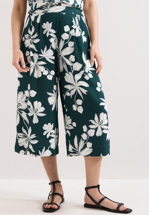 Vrouw met donkergroene, wijde cropped broek met grote witte bloemenprints en zwarte sandalen met bandjes en platte zolen.