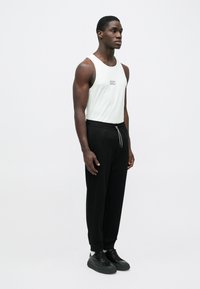 Λευκό αμάνικο τοπ με μικρό λογότυπο, μαύρο παντελόνι με ελαστική μέση και κορδόνι, και μαύρα αθλητικά παπούτσια με παχιά σόλα.