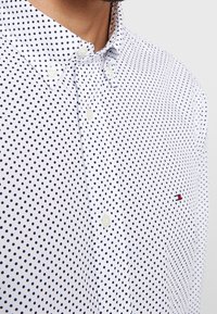 Witte blouse met marineblauwe stippen, voorzien van een klassieke kraag en een klein rood en blauw logo op de linkerborst.