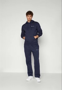 Junger Mann steht mit Händen in den Taschen, trägt einen navyblauen Champion-Hoodie und Sweatpants sowie weiße Sneaker, vor einem schlichten hellen Hintergrund.