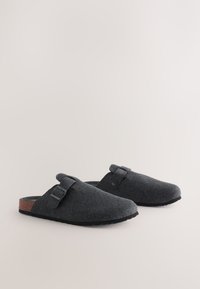 Mules en feutre gris avec bout arrondi, détail de boucle latérale et semelle extérieure en caoutchouc noir contrastant. Surface texturée à l'apparence douce.