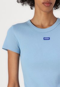 Zbliżenie na osobę noszącą jasnoniebieską, prążkowaną bluzkę z krótkim rękawem i okrągłym dekoltem, z małą niebieską naszywką "HUGO" na piersi.