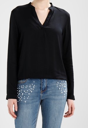 Kvinde iført en sort langærmet V-hals bluse og blå jeans med hvide perlebesætninger på forlommerne.