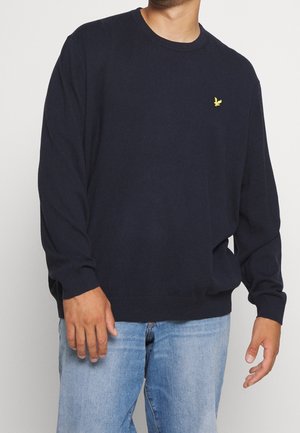 Muž v tmavě modrém svetru s dlouhým rukávem s malým žlutým logem ptáka na hrudi, v kombinaci se světle modrými džíny, stojící na bílém pozadí.