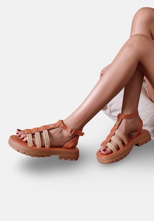 Jambes croisées, portant des sandales beige avec des semelles épaisses, des brides tressées et des boucles à la cheville, reposant sur une surface claire.