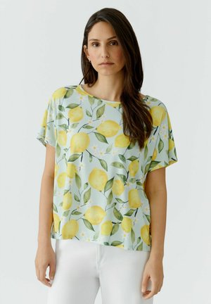 Frau mit langen braunen Haaren, die ein locker sitzendes, kurzärmeliges mintgrünes Hemd mit gelbem Zitronen- und grünem Blattmuster und weiße Hosen trägt.