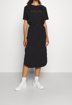 Frau trägt ein schwarzes Midikleid mit kurzen Ärmeln und dem Schriftzug "DKNY New York" sowie weiße Turnschuhe mit hohen Socken, steht vor einem schlichten Hintergrund.