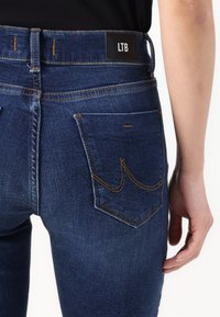Jeans de mezclilla azul oscuro con forma ajustada, que presentan una etiqueta negra de LTB en la cintura, costuras doradas y bolsillos traseros estándar.