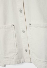 Primo piano della parte anteriore di una giacca di denim color avorio con due grandi tasche applicate e bottoni automatici argento lungo il centro.