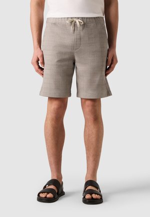 Mann trägt beige karierte Shorts mit Tunnelzug und schwarze Sandalen, steht vor einem einfarbigen hellen Hintergrund.