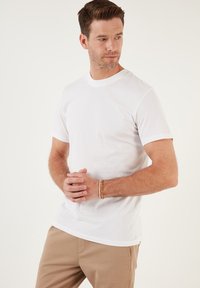 T-shirt bianco in cotone con maniche corte, colletto arrotondato e una texture liscia. Design semplice, indossato con pantaloni khaki e un braccialetto di perline.