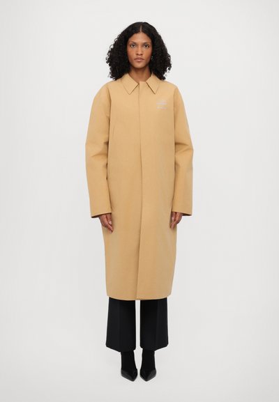 Beige dlouhý kabát s pevným límcem, oversized střihem a bočními kapsami. Nesený přes černé široké kalhoty a špičaté černé boty.