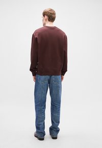 Μπορντό φούτερ με μακριά μανίκια, ριμπ μανσέτες και μέση. Μπλε τζιν με χαλαρή εφαρμογή και δύο πίσω τσέπες. Απλό φόντο.