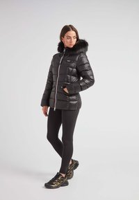 Czarna kurtka puchowa z kapturem z sztucznego futra, z zamkiem błyskawicznym i kieszeniami bocznymi. Połączona z czarnymi legginsami i czarnymi butami sportowymi.