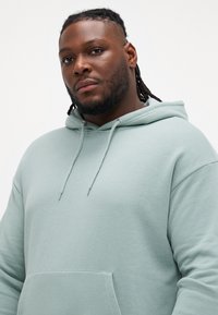 Lichtgroene oversized hoodie van zacht stof met een voorkantzak, een trekkoordkap en ribgebreide mouwen. Soepele textuur en casual ontwerp.
