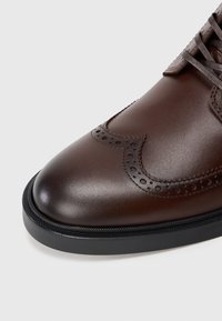 Pantofii de piele maro cu vârful rotunjit, perforații decorative pe partea superioară și o talpă din cauciuc negru. Finisaj texturat.