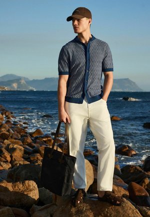 Man wearing patterned blue short-sleeve shirt, beige pants, brown sandals, and cap stands on rocky shore holding black tote bag by the sea.