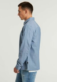 Camicia azzurra chiara con maniche lunghe, taglio dritto, spacco laterale e una texture morbida. Indossata con jeans azzurri chiari.