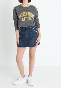 Sudadera gris con el texto "Abercrombie & Fitch" en amarillo, combinada con una falda vaquera con bajo deshilachado y detalles desgastados, y zapatillas blancas.